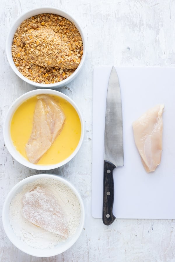 Three bowls full of pecan crust ingredients next to a chicken breast on a cutting board.