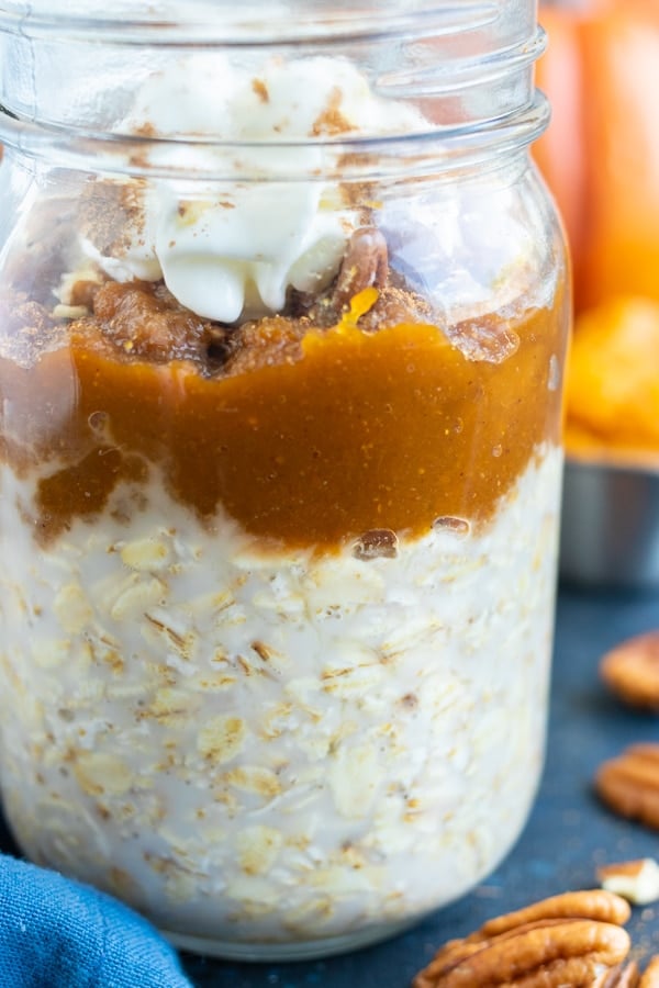 A close-up of a glass mason jar full of overnight oats, pumpkin pie spice puree, toasted pecans, and whipped cream.