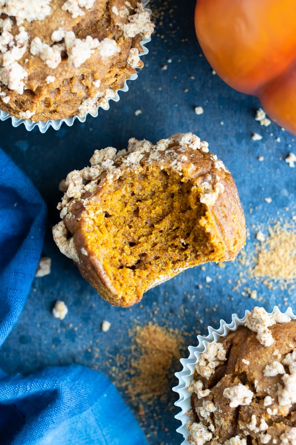 A pumpkin muffin with a bite taken out of it next to two other pumpkin streusel muffins.