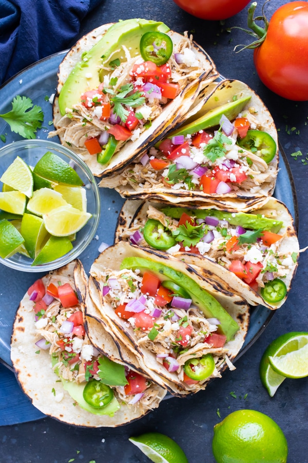 A plate full of shredded chicken tacos with tomatoes, avocado, cheese, and lime wedges.