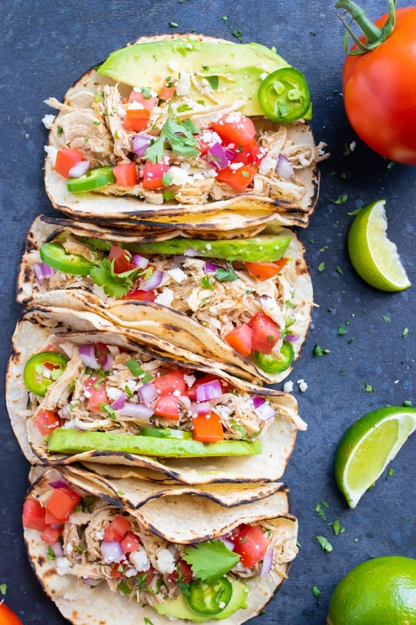 Four shredded chicken tacos in a row with tomatoes an avocado next to lime wedges.