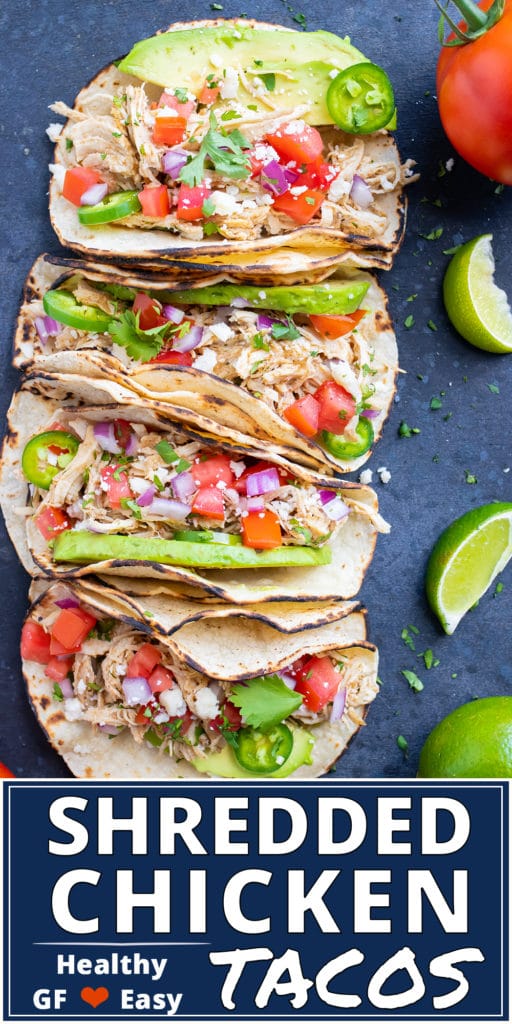 Four shredded chicken tacos in a row with tomatoes an avocado next to lime wedges.
