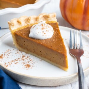 A slice of the best homemade pumpkin pie recipe with whipped cream and cinnamon sticks next to it.