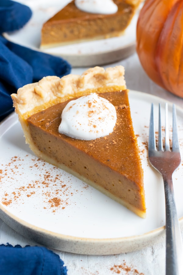 A slice of the best pumpkin pie recipe from scratch with whipped cream and cinnamon sticks next to it.
