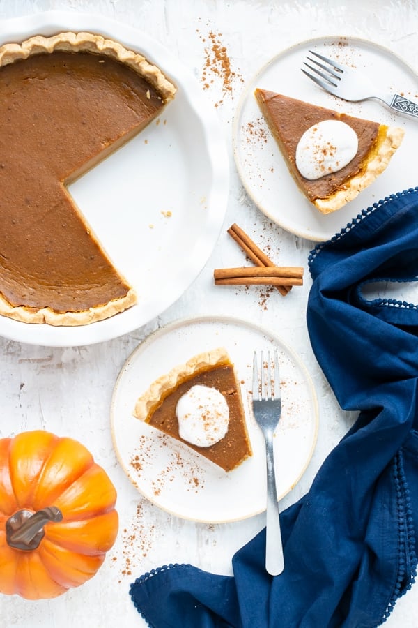 Two dessert plates with slices of the best pumpkin pie recipe next to a blue napkin and a 9-inch pie dish.
