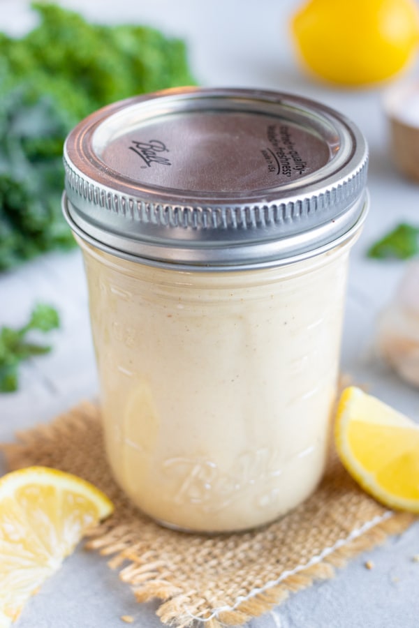 A jar of lemon tahini salad dressing sits on burlap next to lemon wedges.