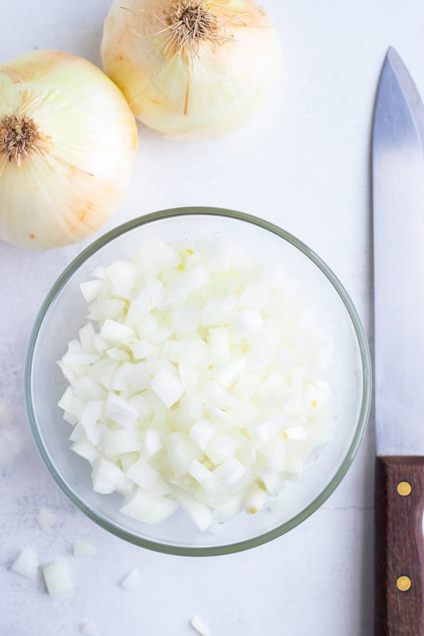 A bowl of diced onion sits on a white cutting board next to a sharp chef knife.