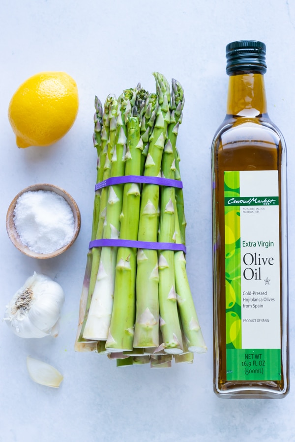 A bunch of asparagus, olive oil, lemon, salt, and garlic as the ingredients for a roasted asparagus recipe.