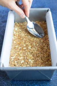 A spoon pressing down the oatmeal crumble to form the base of Samoa bars in a loaf pan.