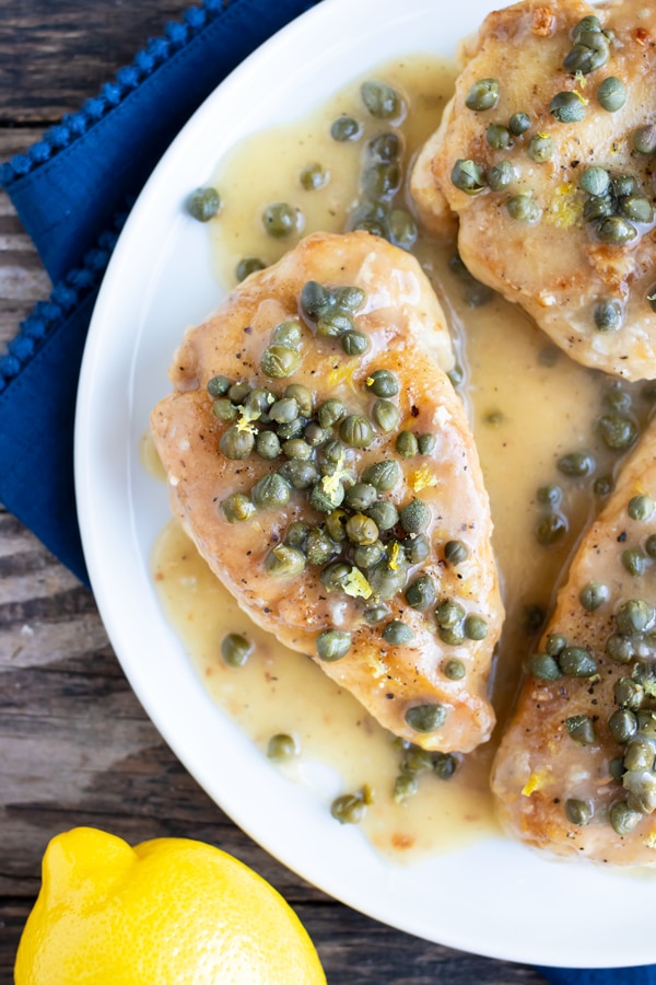 Healthy chicken piccata on a white plate with capers and sauce next to a lemon.