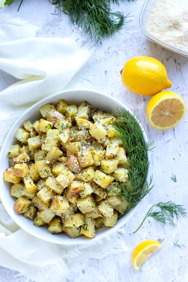 A bowl full of a potato side dish recipe next to lemons, Parmesan cheese, and fresh dill.