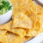 Homemade tortilla chips on a white plate next to a bowl of guacamole.