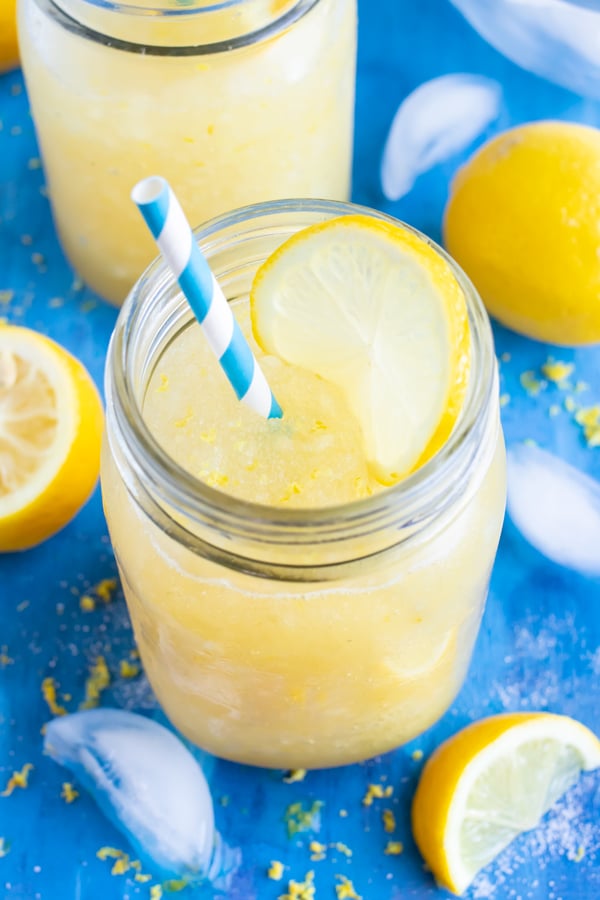 A glass full of a healthy frozen lemonade recipe with a lemon slice in it surrounded by ice cubes.