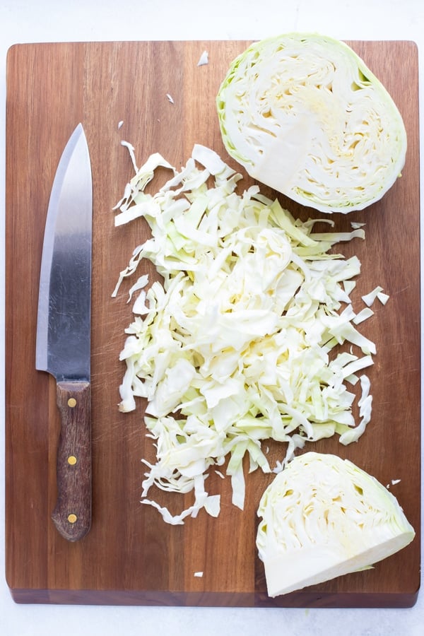 A wooden cutting board with a knife, cabbage wedge, and shredded slices.