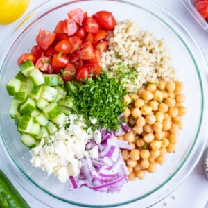 A large mixing bowl with pearl couscous, garbanzo beans, tomatoes, cucumbers, and chopped parsley.