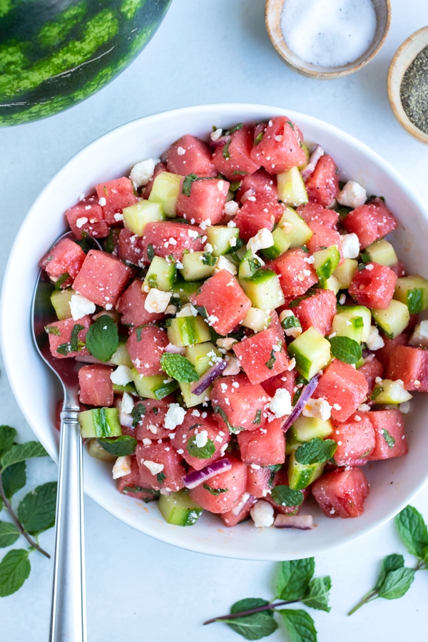 Watermelon salad with feta cheese crumbles, fresh mint, and cucumbers with a silver serving spoon.