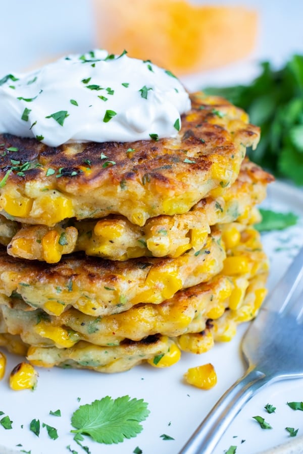 A stack of cheesy corn fritters with a dollop of sour cream and cilantro in the background.