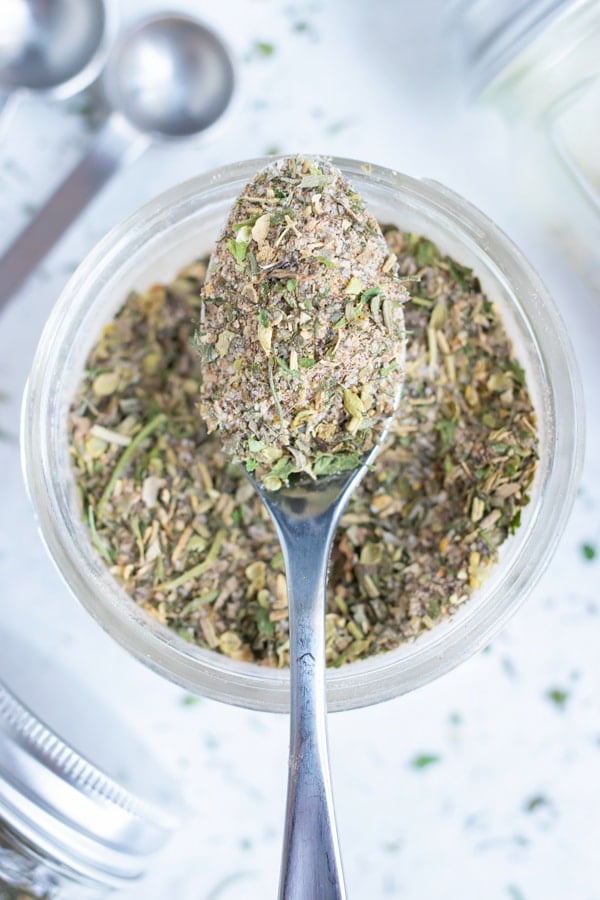 A metal spoon lifts up some Greek seasoning with dried herbs and spices from a glass jar.