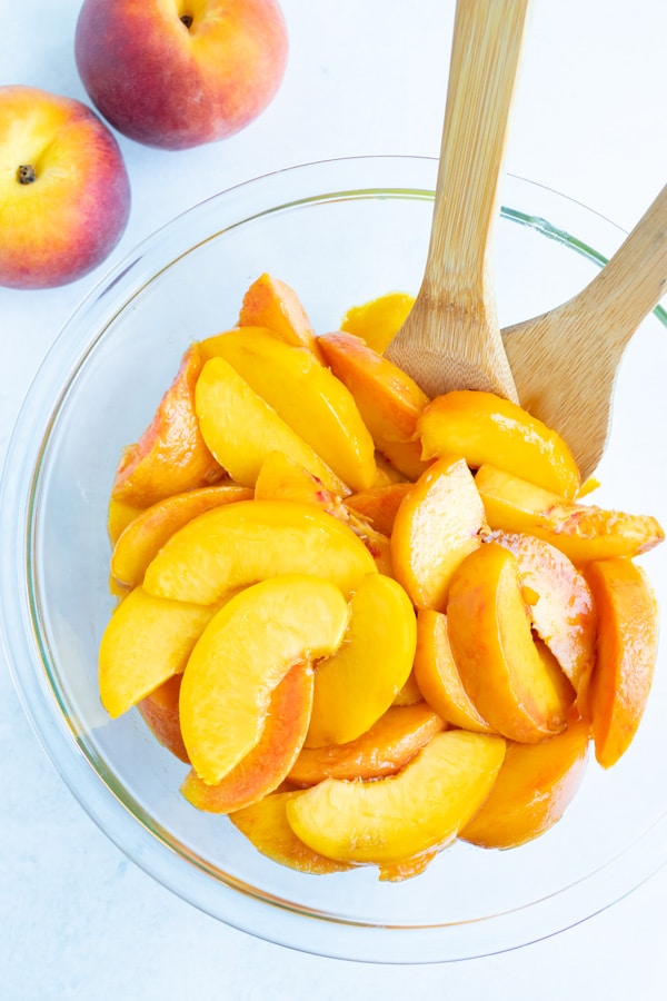Peeled, sliced peaches are sitting in a bowl with wooden spoons on the counter in preparation for a recipe.