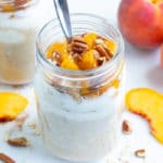Two mason jars full of peach oatmeal are on the counter with peaches and pecans.