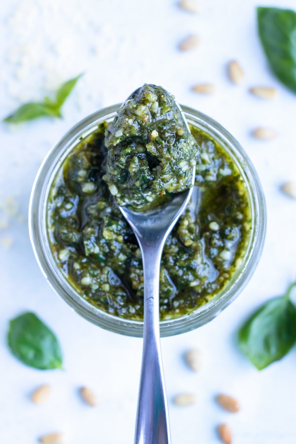A spoon holds basil pesto sauce from a mason jar, with fresh basil leaves on the counter.