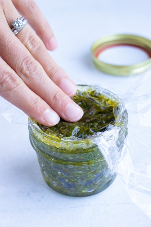 Storing leftover basil pesto in an airtight container covered with a piece of plastic wrap.