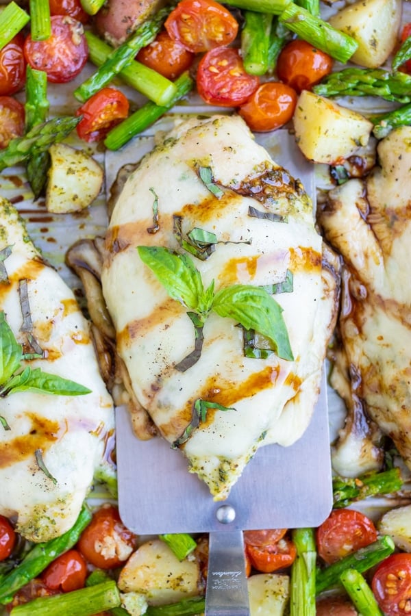 Cubed potatoes, sliced tomatoes and asparagus covered in pesto are cooked together with caprese chicken topped with melted mozzarella on a sheet pan and then drizzled with balsamic vinegar and sprinkled with fresh basil leaves.