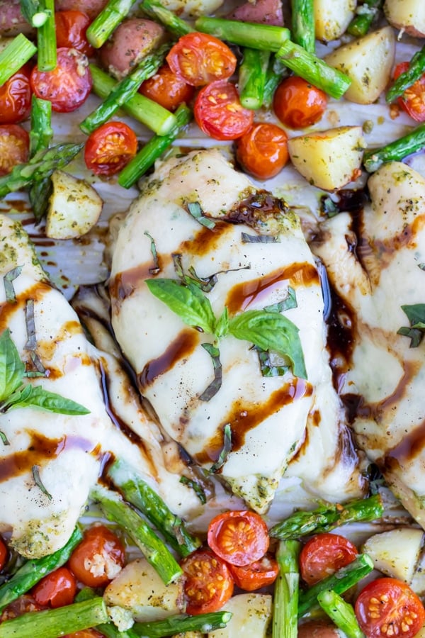 Baked chicken caprese covered with mozzarella cheese and pesto is cooked on a large baking sheet with potatoes, asparagus, and tomatoes with a balsamic glaze drizzle over top.
