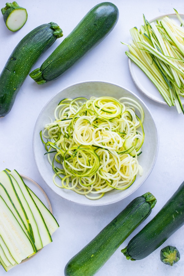 A pile of spiralized zucchini in a bowl on the counter with sliced and thin zucchini on a plate nearby.