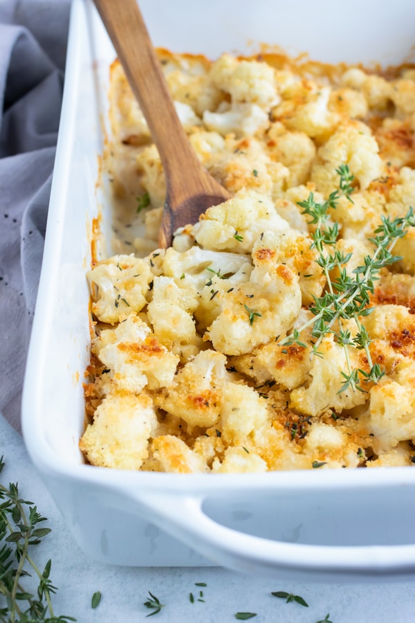 Keto cauliflower gratin is served with a wooden spoon from a casserole dish.