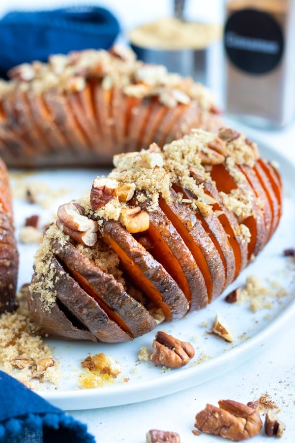 Hasselback sweet potato recipe is served on a white plate and topped with toasted pecans, brown sugar, and cinnamon.