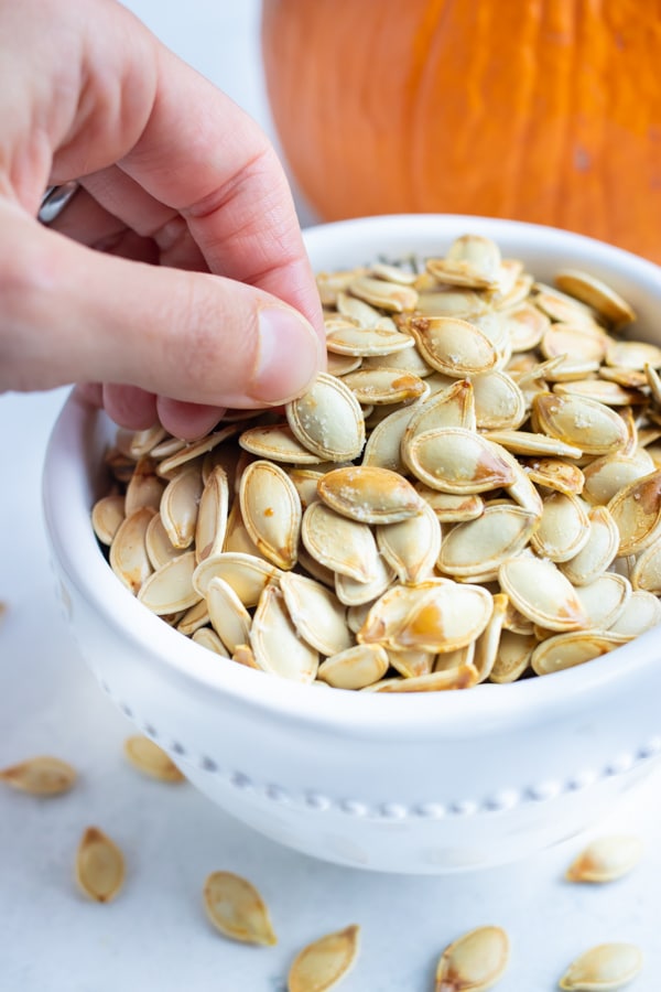 Pumpkin seeds in a white bowl are picked up for a snack.