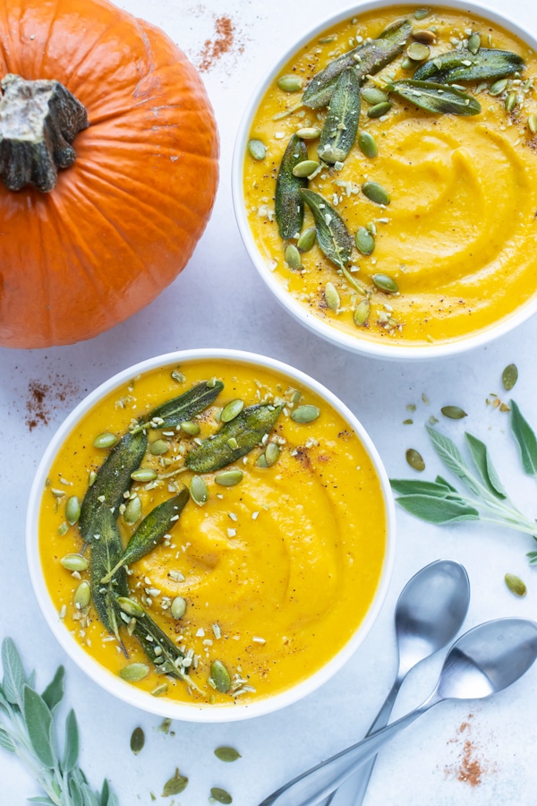 Dairy-free pumpkin soup is served in white bowls and topped with herbs and pumpkin seeds.