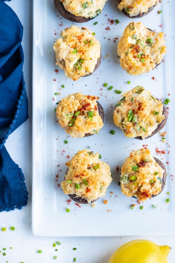 Crab stuffed mushrooms are served on a white platter with chives and paprika.