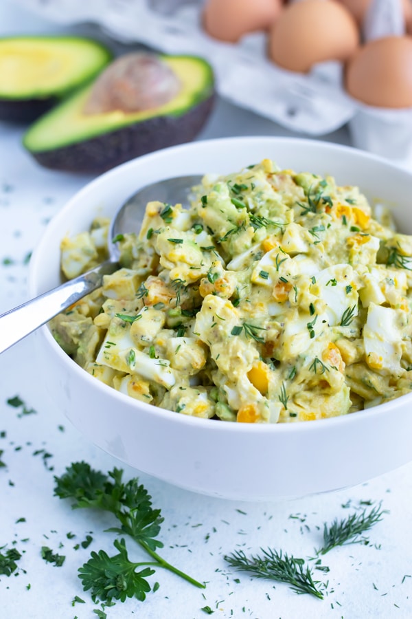 Flavorful avocado egg salad is dished with a spoon from a white bowl.