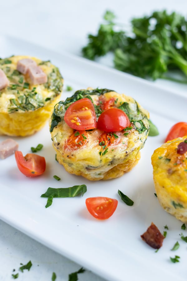 Three breakfast egg muffins are served one a plate with fresh tomatoes and herbs.