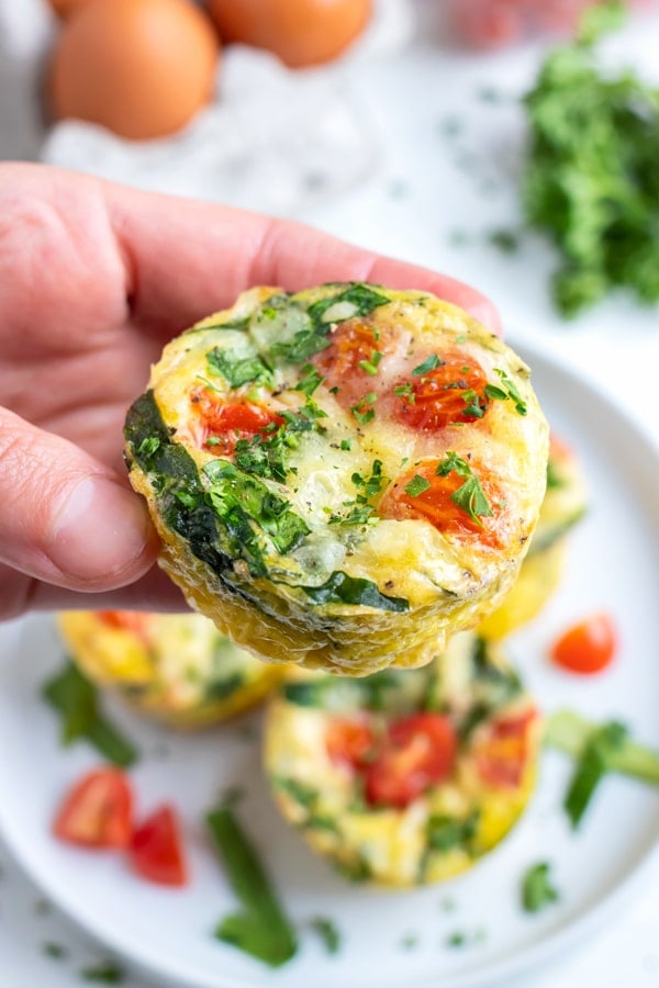 A hand is holding an egg cup full of spinach, tomatoes, and cheese.
