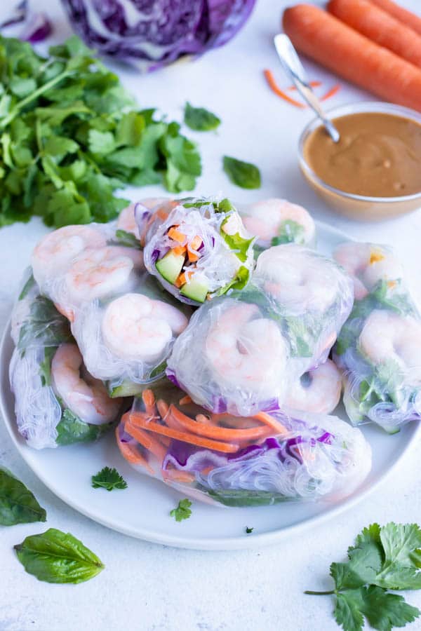 A pile of spring rolls are served with peanut sauce, carrots, cabbage, and basil around them.