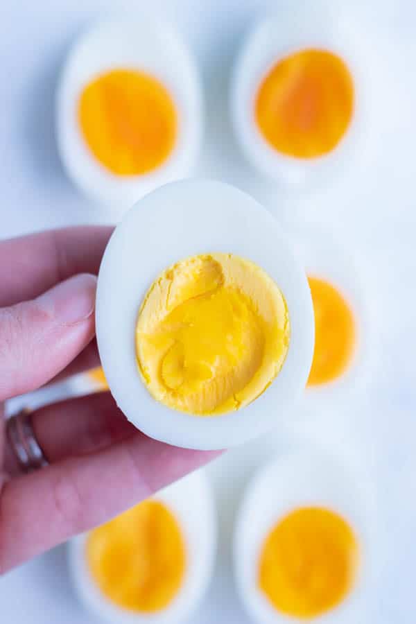 A hard-boiled egg is held by a hand with more eggs behind it.