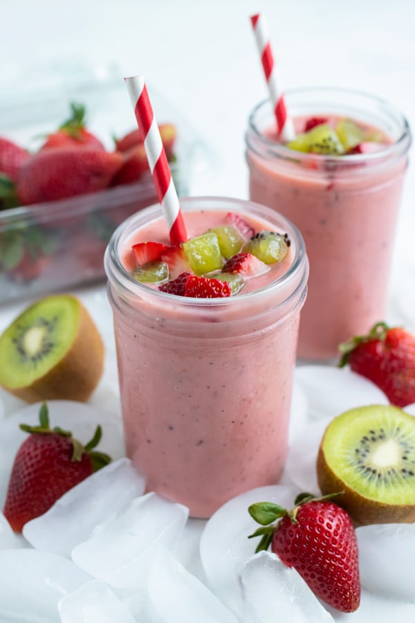 Two cups of strawberry smoothies are set on the counter next to fresh strawberries and kiwis.