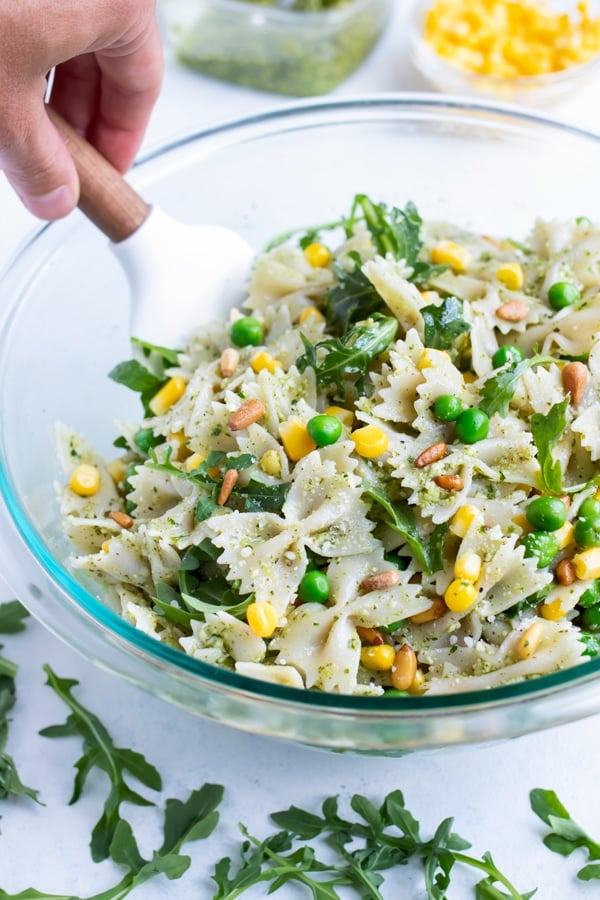 Pesto pasta salad is mixed in a glass bowl with a rubber spatula.