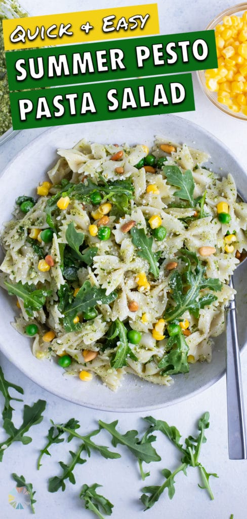A bowl of summer pasta salad is shown on the counter next to pesto and corn.
