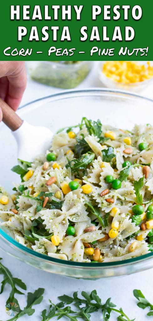 Pesto pasta salad is mixed in a glass bowl with a rubber spatula.