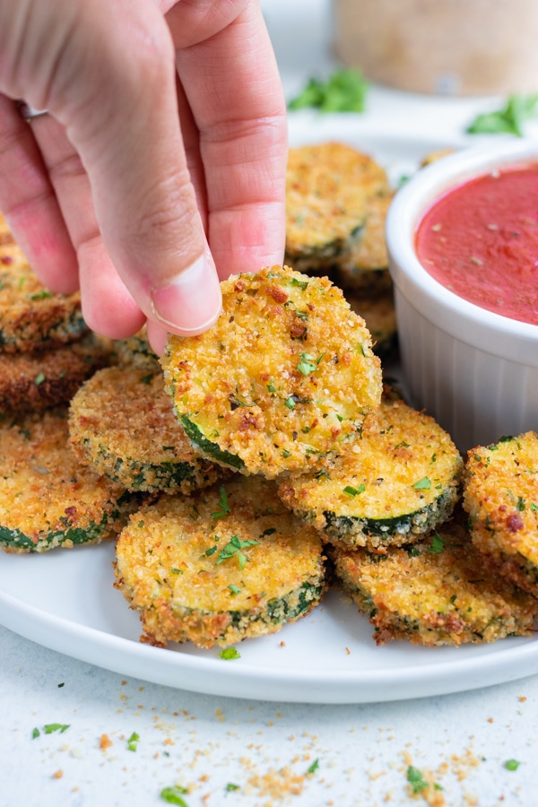 A hand is used to enjoy air fryer zucchini chips from a plate.