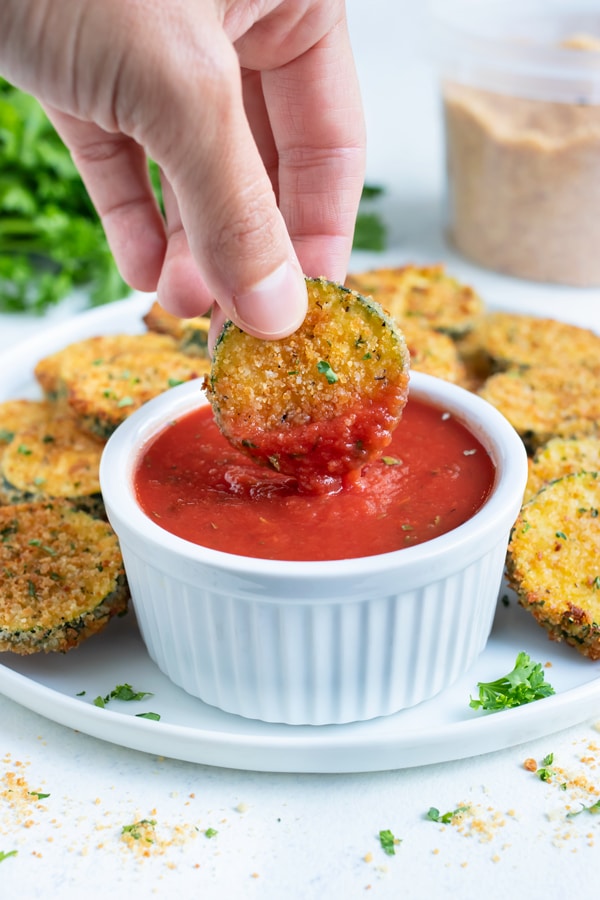 An air fryer zucchini chip is lifted up and dipped into marinara.