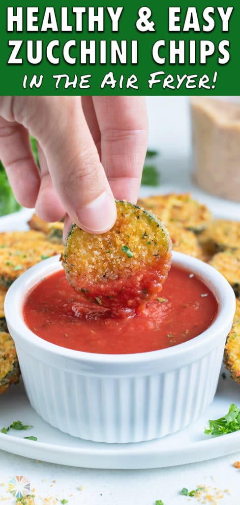An air fryer zucchini chip is lifted up and dipped into ketchup.