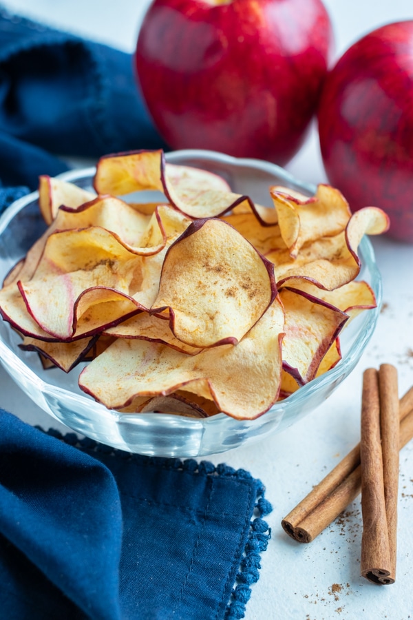 A bowl of apple chips is set next to sticks of cinnamon.