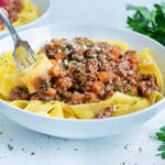 A fork is shown twisting noodles with bolognese sauce from a bowl.