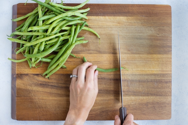 A green bean is cut with a knife on a cutting board.