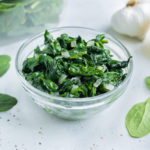 A bowl of sautéed spinach rests on the counter by fresh spinach.